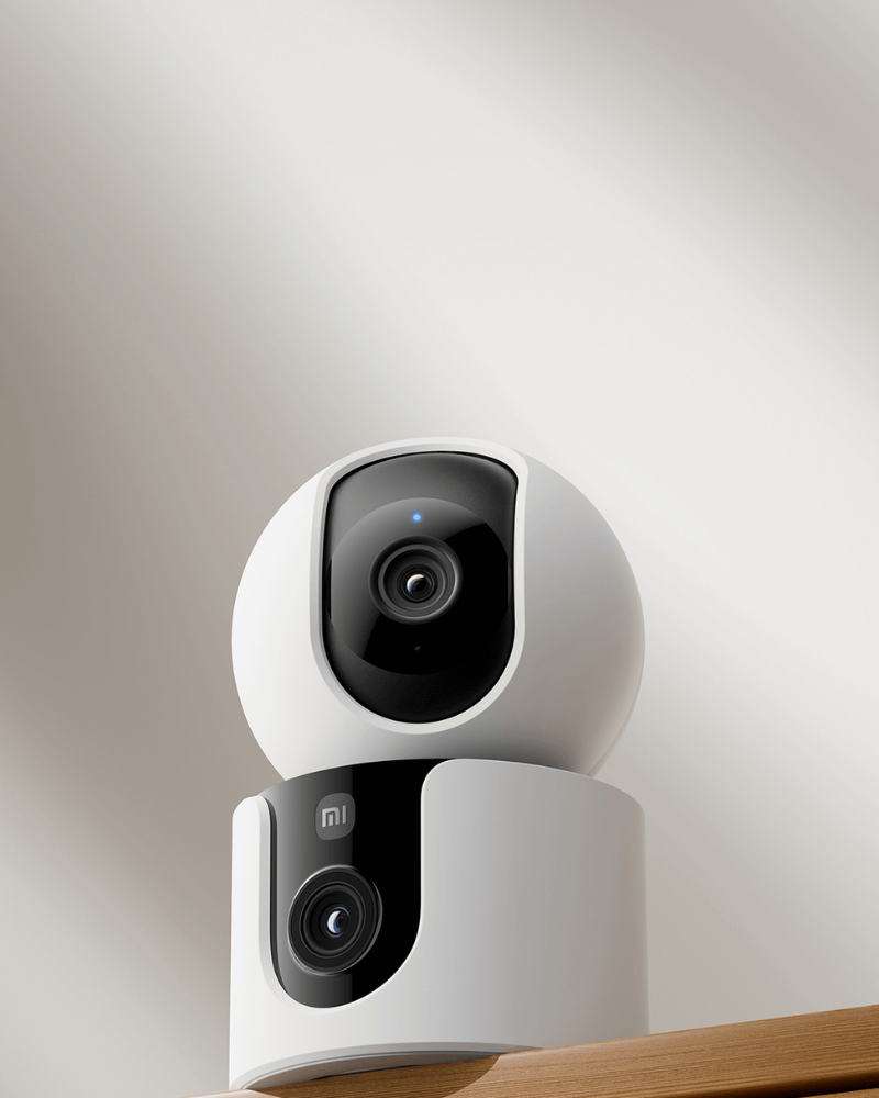 A close-up product image of a Xiaomi smart security camera placed on a wooden surface, highlighting its dual-lens design for home monitoring.