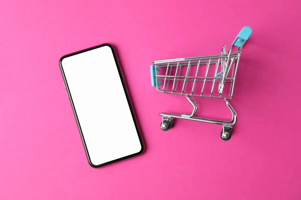 Smartphone with a blank screen next to a shopping cart on pink. Find your ideal AI phone with our guide.