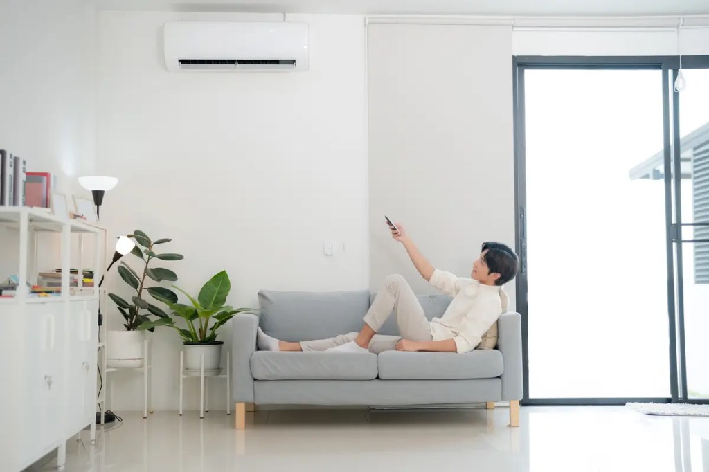 A young man relaxing on a gray sofa in a minimalist living room, using a remote control to adjust a budget-friendly wall-mounted air conditioner during a heatwave.